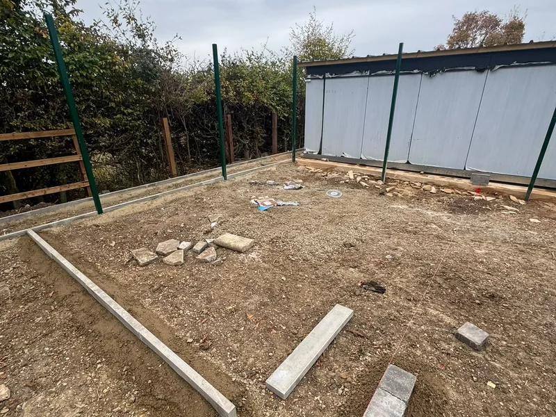 Fenced site compound with levelled ground and concrete kerbs beside a clad building