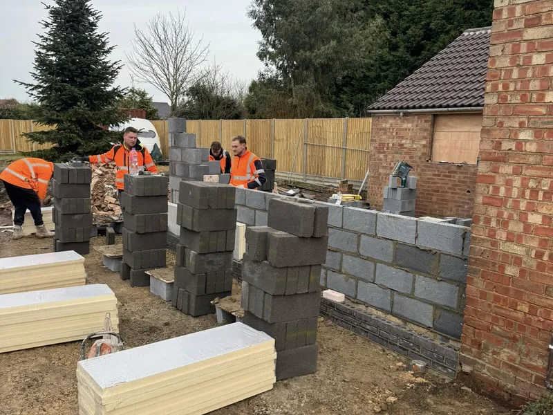 Two operatives laying blockwork with concrete lintels and insulation boards stacked on site