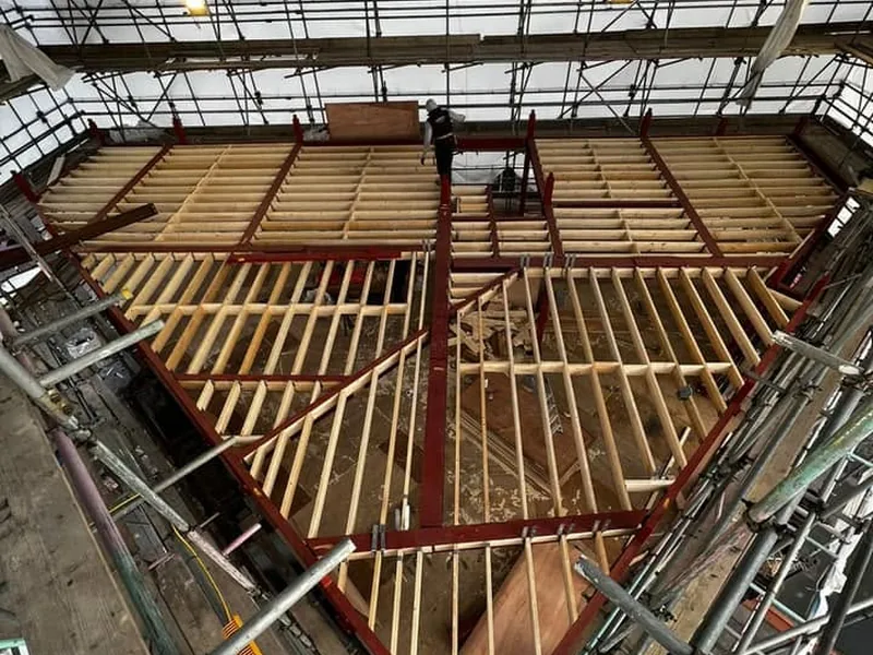 New timber floor joists spanning red steel beams at roof level under a temporary scaffold roof