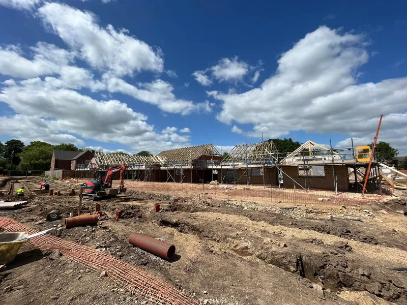 Drainage trenches and an excavator on a new build site with roof timbers going up behind