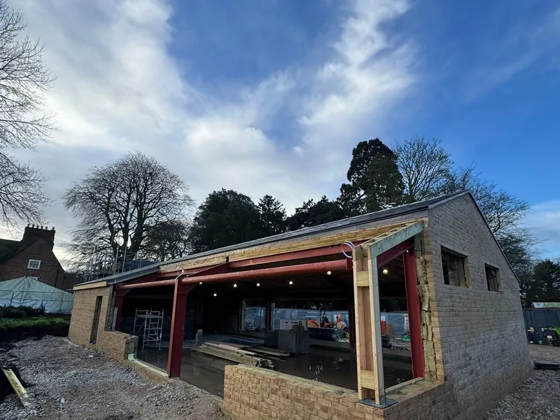 Single storey building under construction with a red steel frame, timber posts and open glazed bays