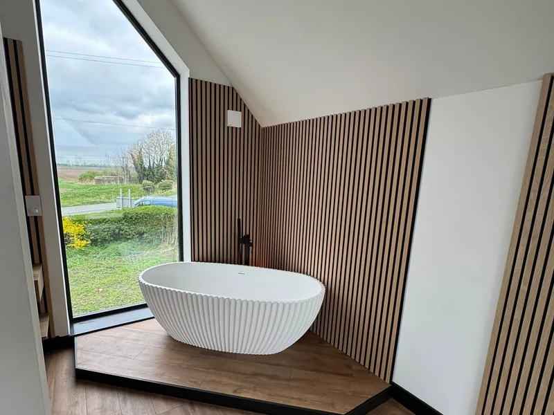 Freestanding bath in a room lined with slatted timber wall panelling beside a tall window